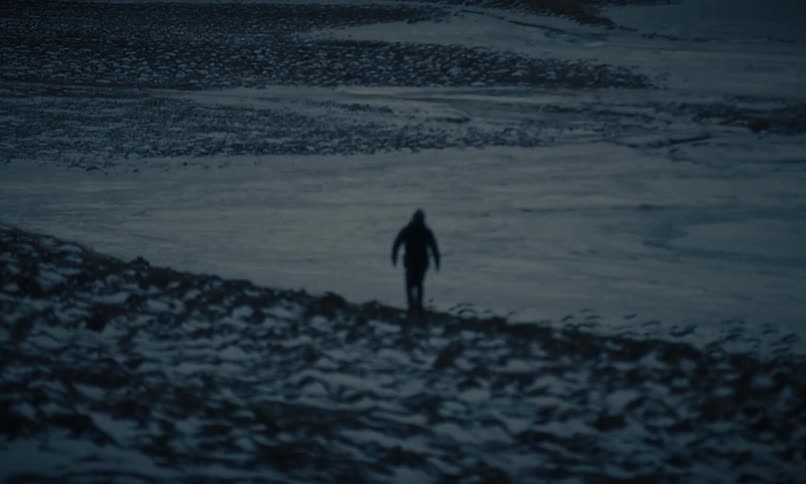 A blurry, silhouetted figure walks across a desolate, dark landscape before stopping to look out at the emptiness