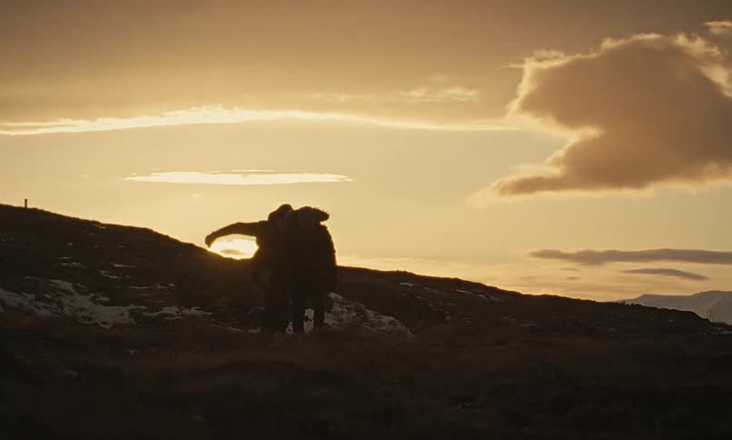 A silhouetted couple playfully hugs, dances, and gives a piggyback ride while hiking on a mountain at golden hour