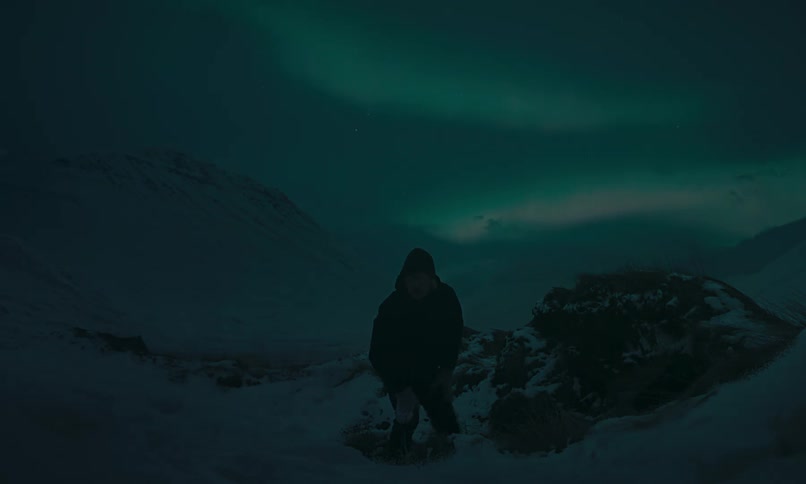 A man stands up in a snowy mountain landscape at night to watch the beautiful Aurora Borealis light up the sky