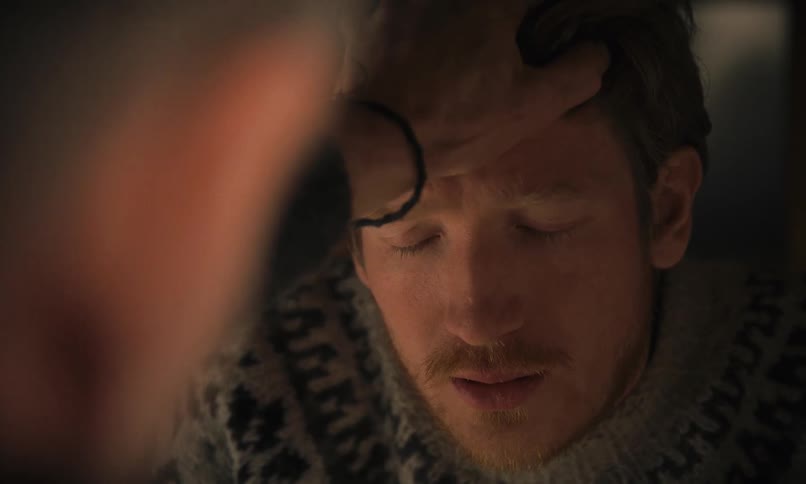 An emotional close-up of a man receiving a comforting hand on his forehead in a dimly lit room