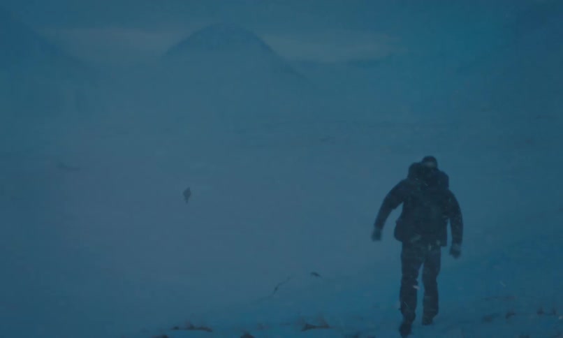 A mountaineer struggles through a blizzard in a desolate, snowy mountain valley, following another person in the distance