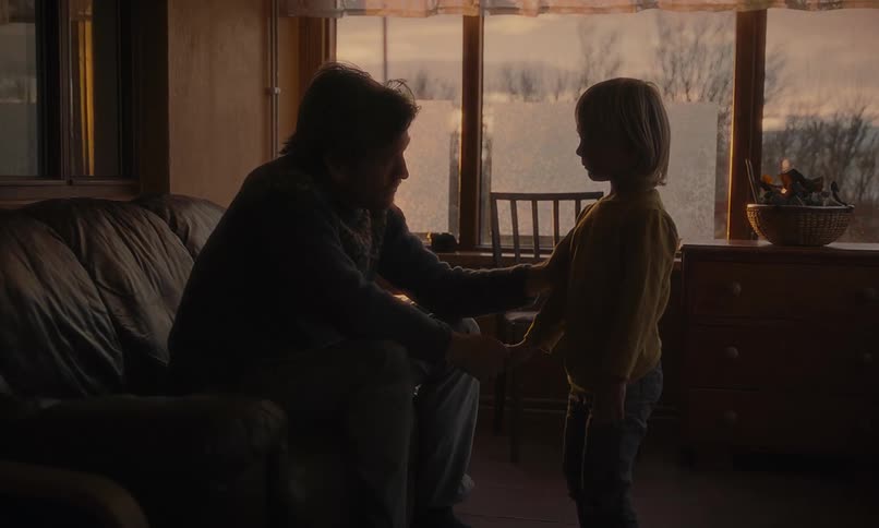 A heartbroken father sits on a couch in a dimly lit room as his young son walks away after a sad farewell