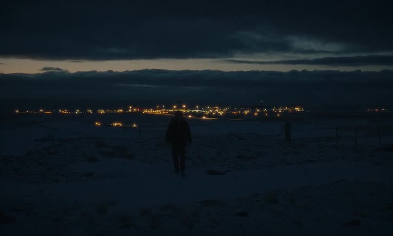 A lone person walks through a dark, snowy field at twilight, with the warm lights of a distant village on the horizon