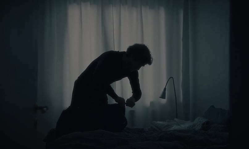 A man silhouetted by a window unpacks his bag in a dark and moody bedroom