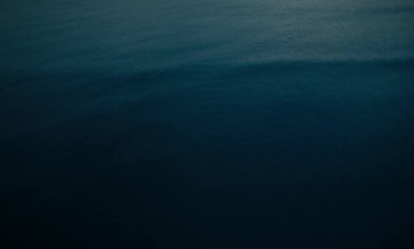 Top-down aerial view of a person in a bright orange jumpsuit lying in the dark, foamy surf on a black sand beach at night