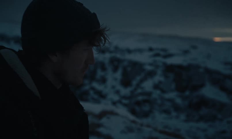 A lone hiker walks through a desolate, snowy canyon alongside a frozen river at twilight, stopping to take in the vast landscape