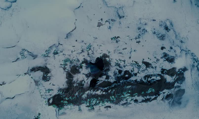 An aerial top-down view shows a person performing an abstract dance with a colorful, flowing fabric in a desolate, snowy landscape