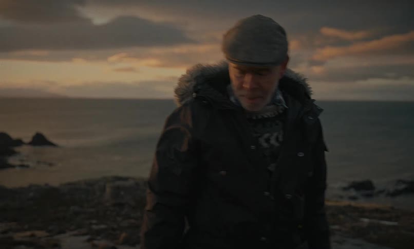 A solitary man in a winter coat thoughtfully walks along a rugged, snowy coastline at sunset towards a distant lighthouse