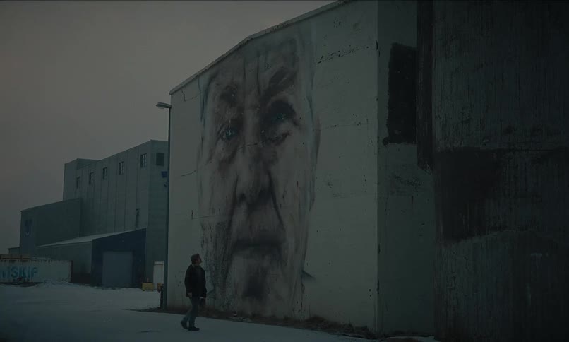A lone figure walks through a desolate, snowy industrial area past a striking mural of an old man's face