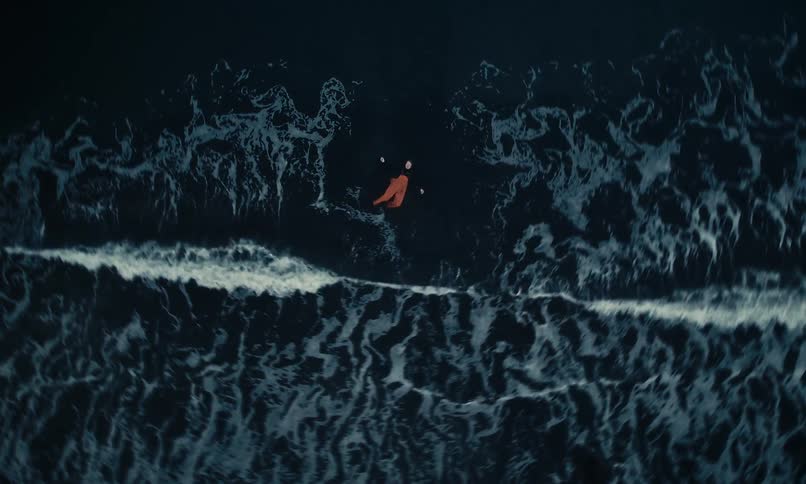 A striking aerial cinemagraph shows a person in orange pants floating calmly on the dark ocean as waves roll over a black sand beach