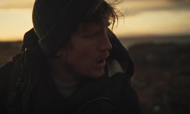A weary young traveler with a backpack looks up in shock and alarm in a desolate, windswept landscape at sunset