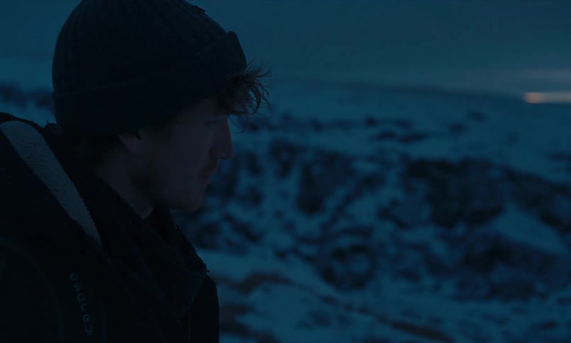 A lone hiker walks through a vast, snowy canyon in Iceland at twilight, followed by another man who continues the journey through the desolate landscape