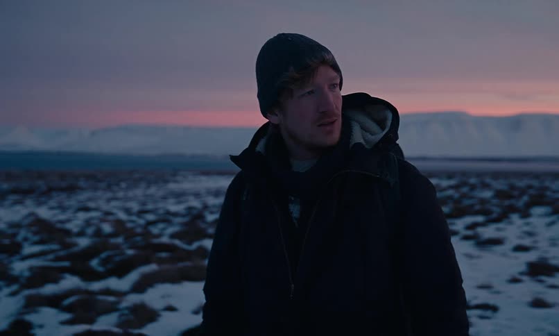 A man wearing a winter coat and beanie stands in a vast, snowy field, watching the sunset over the mountains