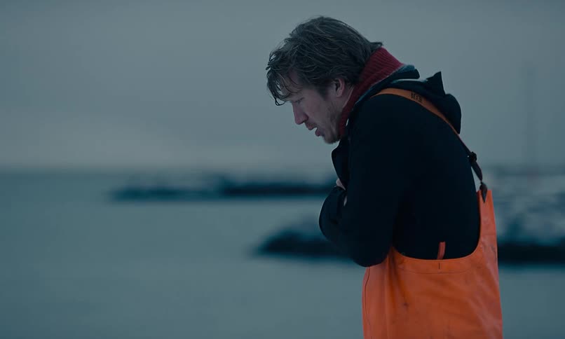 A cold fisherman in orange overalls shivers as he walks through a snowy harbor on a bleak winter day