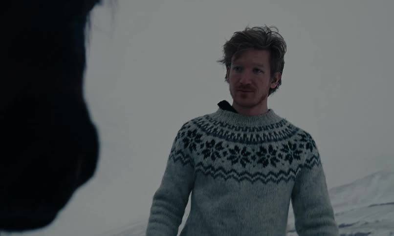 A man in a traditional Nordic sweater gently approaches a horse in a snowy, mountainous landscape