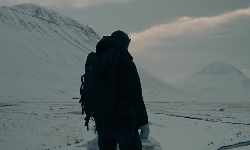 A lone hiker stands and contemplates a vast, snowy mountain landscape on a cold, overcast day