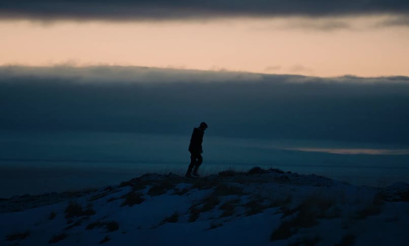 Slow motion shot of a silhouetted man walking along a snowy hill at dusk with the ocean in the background