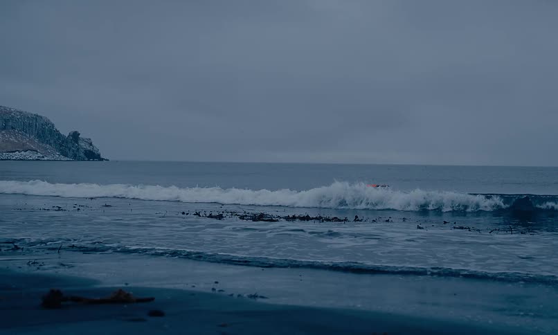 Slow-motion waves roll onto a desolate, dark sand beach on a cold, overcast day