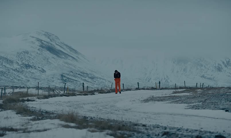 A man in bright orange pants walks through a vast, cold, and snowy mountain landscape