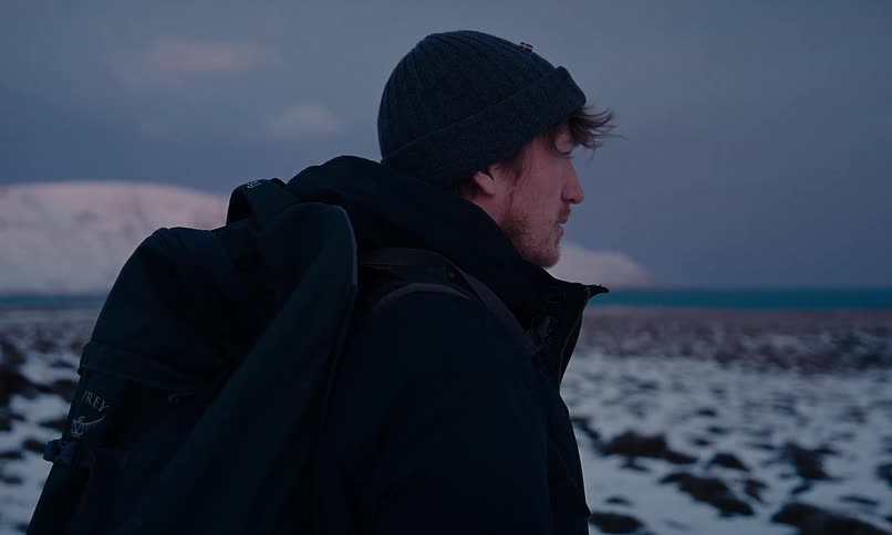 A male adventurer wearing a backpack and winter gear stands in a vast, snowy landscape, looking out towards the sea at dusk