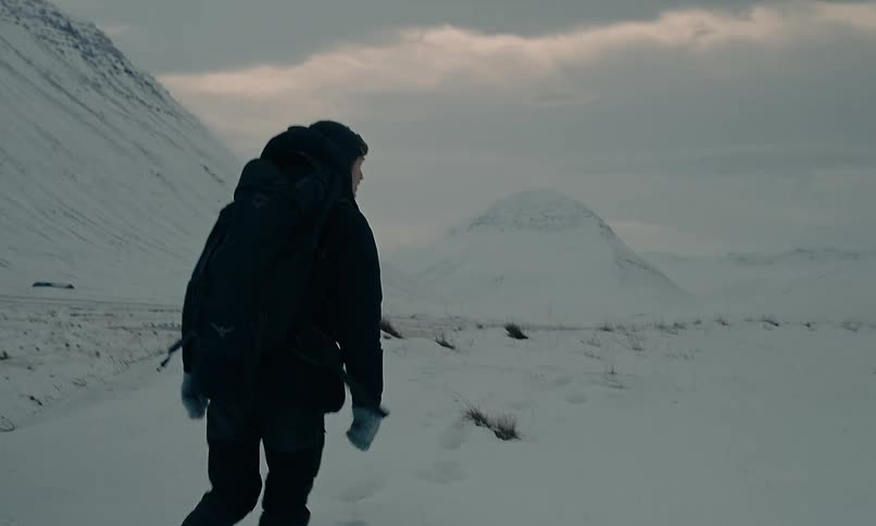 A lone explorer treks in slow motion through a vast and desolate snow-covered valley surrounded by mountains