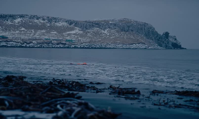 A person wearing a bright orange survival suit floats in the cold ocean as slow-motion waves wash ashore on a winter beach