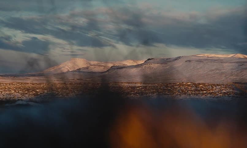 A picturesque, out-of-focus view shows steam rising over a vast, snow-covered Icelandic landscape with mountains lit by the golden hour sun