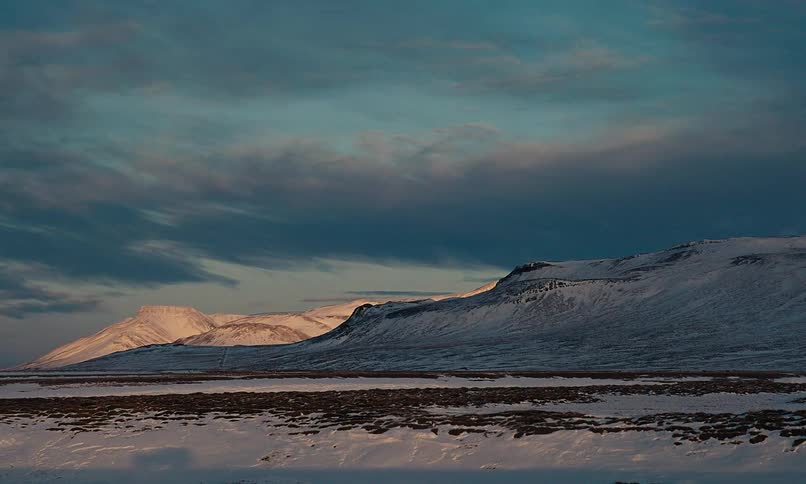 Golden sunset light illuminates the peaks of vast, snow-covered mountains in a serene Icelandic winter landscape