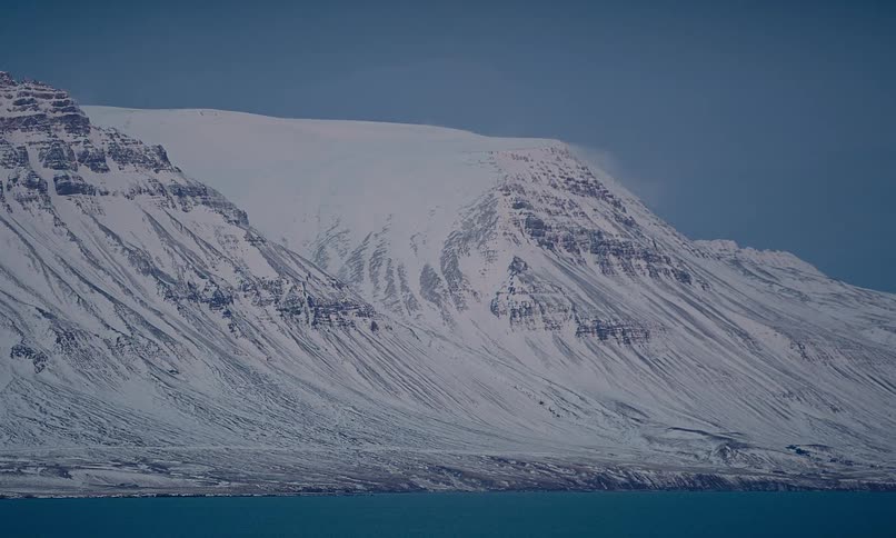A majestic snow-covered mountain range rises from the edge of a calm, deep blue ocean on a cold winter day