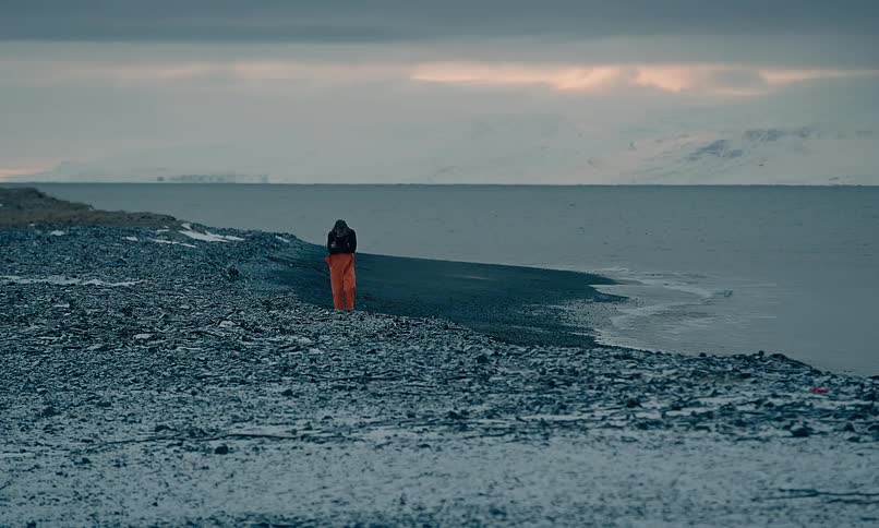 A solitary figure in orange waterproof trousers walks slowly along a vast and desolate frozen shoreline