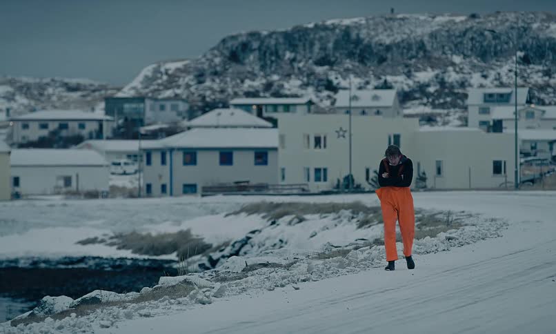 A person in bright orange overalls shivers while walking down a snowy road through a remote coastal village