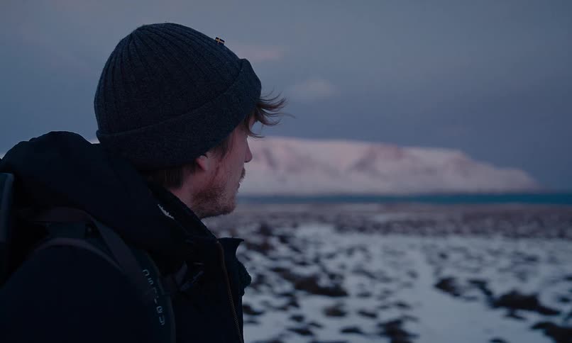 A contemplative man wearing a beanie and backpack observes the vast, snowy landscape during a cold twilight