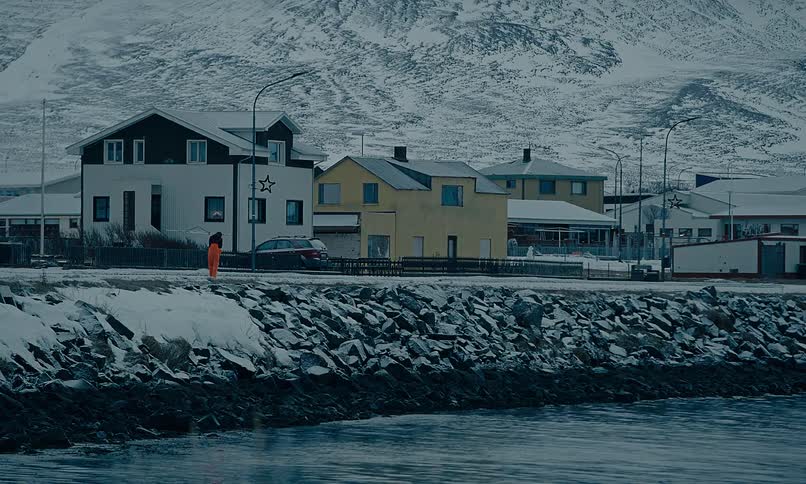 A solitary figure in a bright orange suit stands motionless on a snowy, rocky shoreline in a remote arctic village