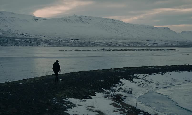 A solitary traveler walks through a remote and snowy Icelandic landscape overlooking a vast fjord and mountains at dusk