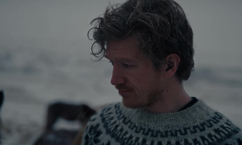 A thoughtful man wearing a cozy wool sweater stands outside as snow gently falls on his hair
