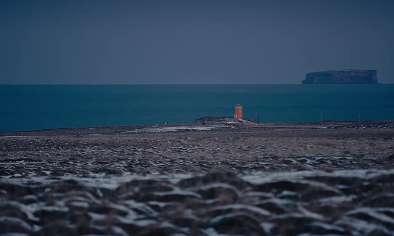 A small orange lighthouse stands on a desolate, snow-dusted coastline overlooking the dark ocean at dusk