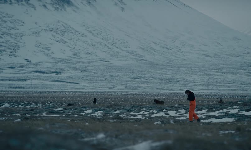 A solitary figure in orange pants walks slowly across a vast, desolate, snowy landscape with mountains in the distance
