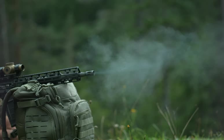 A close-up of a rifle firing in super slow motion, showing the muzzle flash and smoke against a forest backdrop