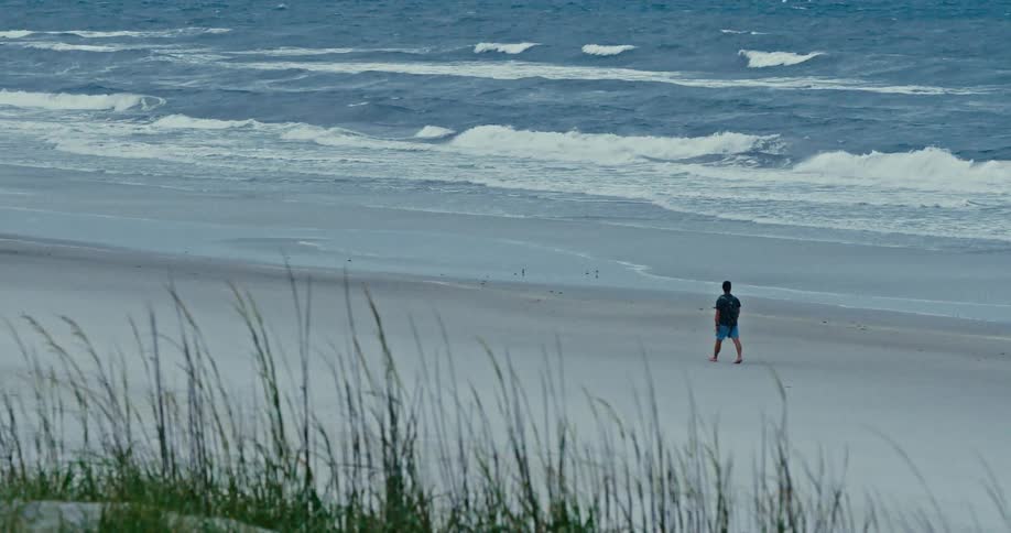 A solitary man takes a contemplative walk along a vast, empty beach as waves roll in from the ocean