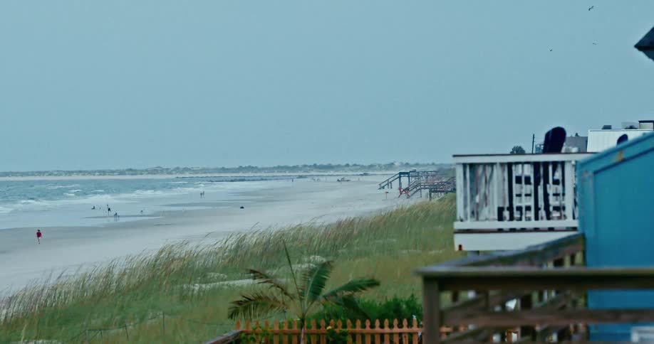 A peaceful view from a deck overlooking a wide sandy beach with distant people walking along the shore on a cloudy, overcast day