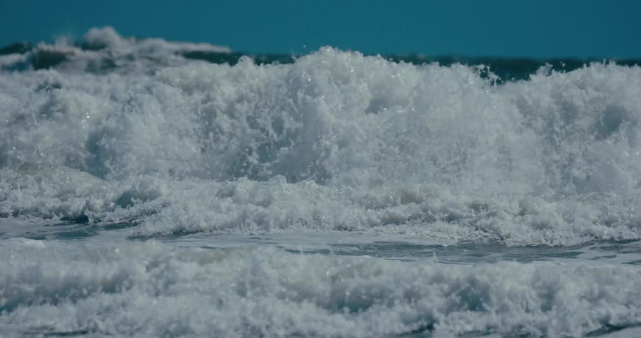 A beautiful emerald green wave forms and breaks into white surf in dramatic slow motion under a clear blue sky