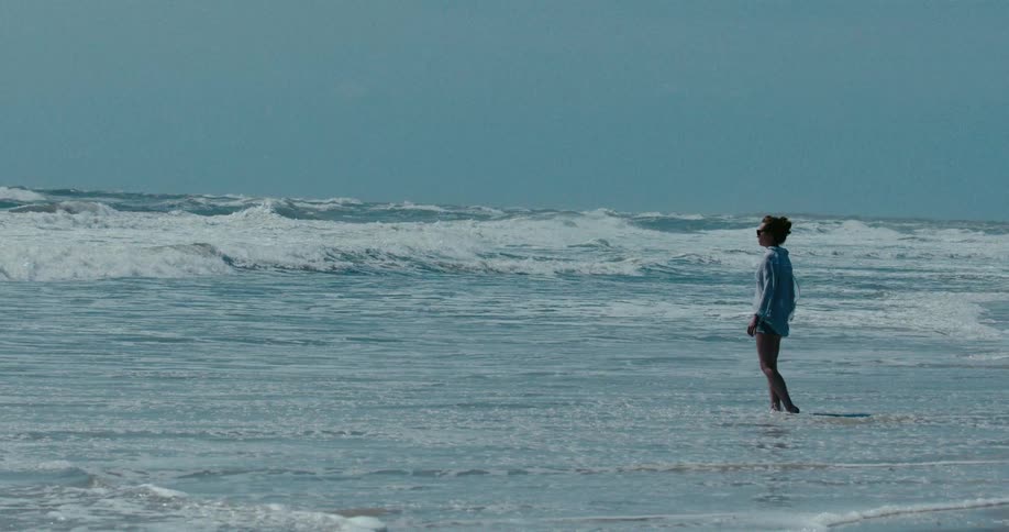 A pensive woman in a blue shirt stands in the shallow ocean surf, watching the waves roll in on a breezy day in slow motion