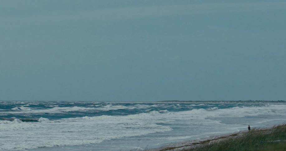 A solitary person observes the vast, rough ocean from the shore on a windy, overcast day