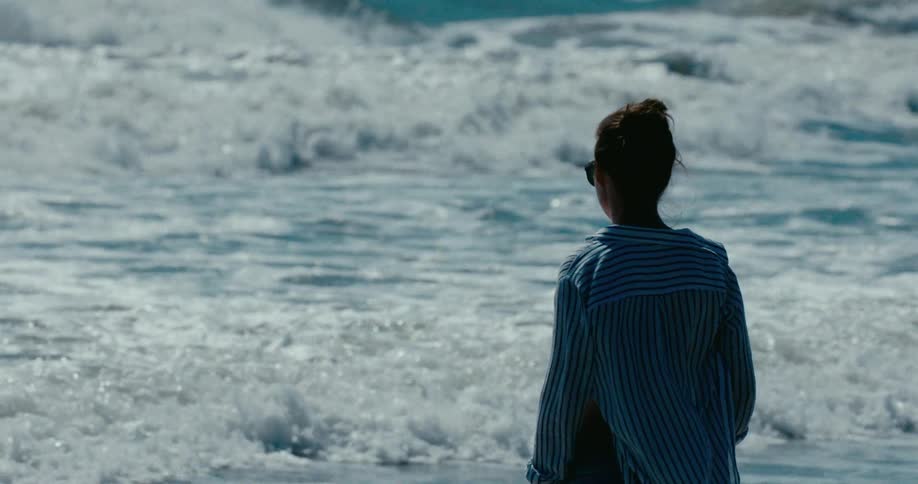 A woman wearing a striped shirt stands on the beach and watches the powerful ocean waves in slow motion