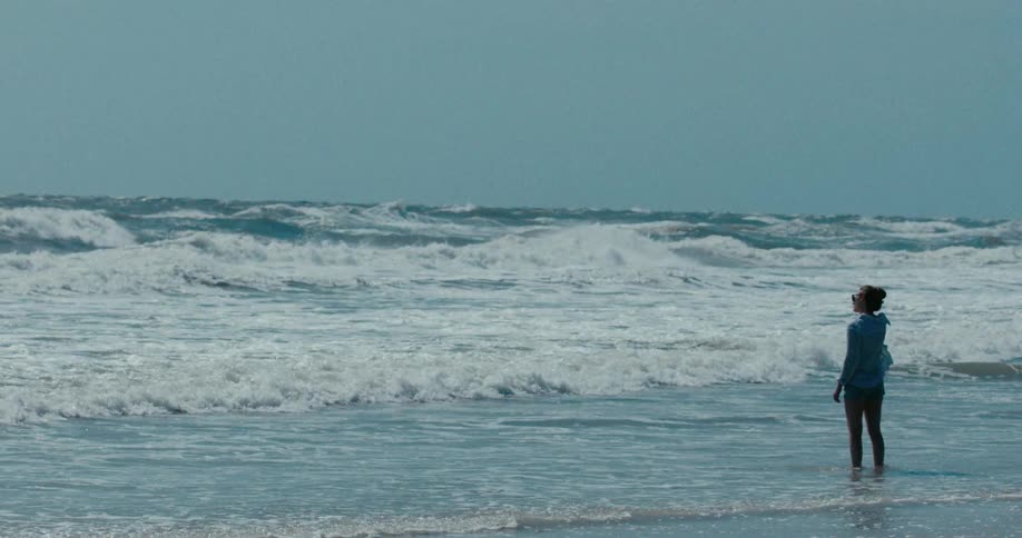 A woman stands in the shallow ocean surf and watches the turbulent waves roll in during a windy day, shot in slow motion