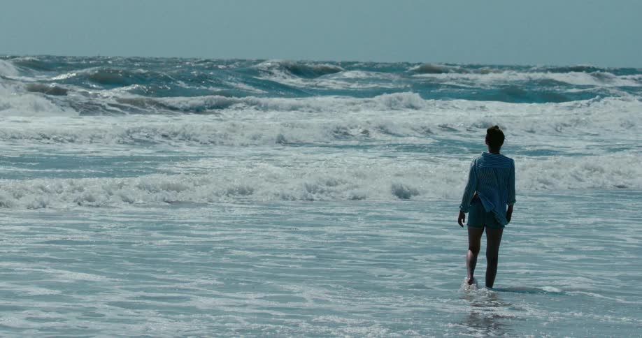 Slow motion shot of a lone woman standing in the rough ocean surf, watching the powerful waves crash on the shore