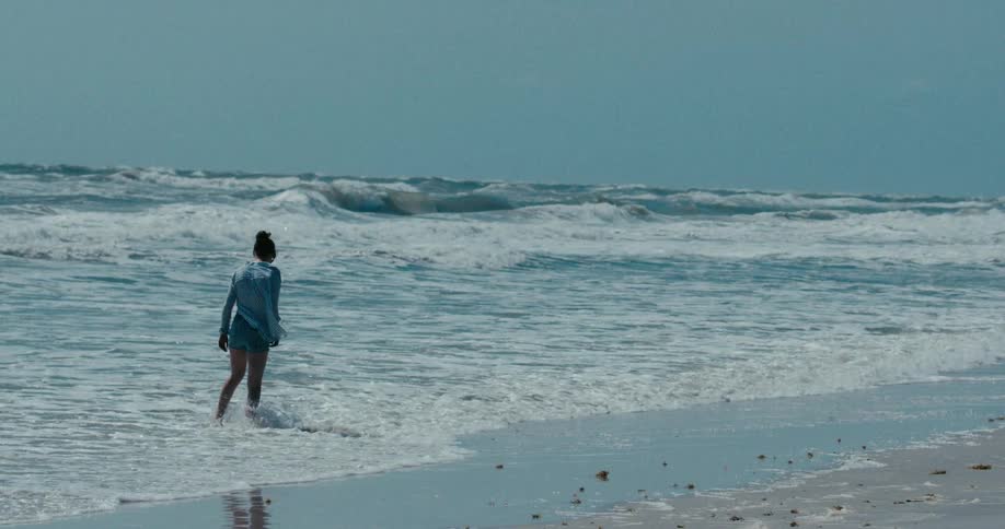 A lone woman walks in slow motion along the shoreline of a choppy ocean, pausing to look out at the waves