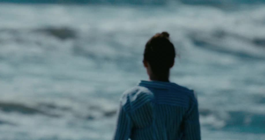 A contemplative woman in a striped shirt stands on the shore, gazing at the rolling ocean waves in slow motion