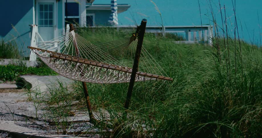 An empty, broken hammock sways peacefully in the tall sea grass behind a beach house on a windy day