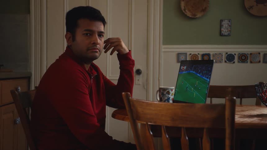 A serious man watches a tense soccer match on a tablet at his kitchen table, turning to the camera with a worried expression